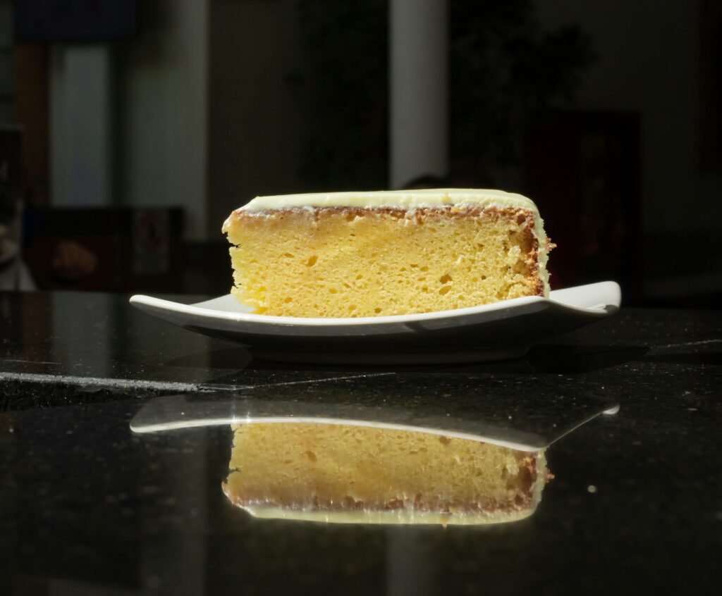 A fresh slice of lemon cake on a white plate, reflecting on a shiny black surface in a dimly lit café.
